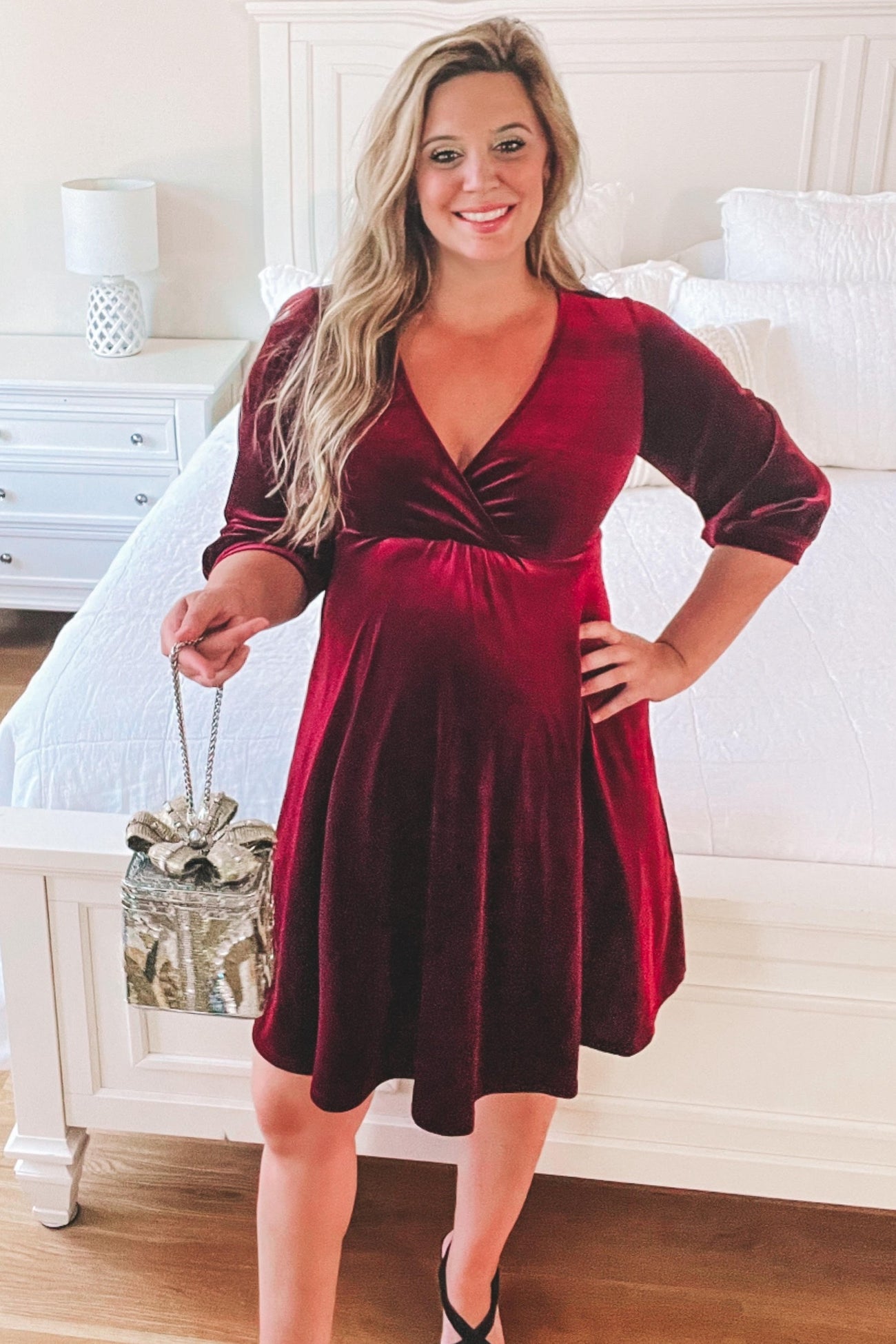 woman wearing a burgundy velvet maternity dress with three-quarter sleeves and holding a sparkly purse, baby bump visible