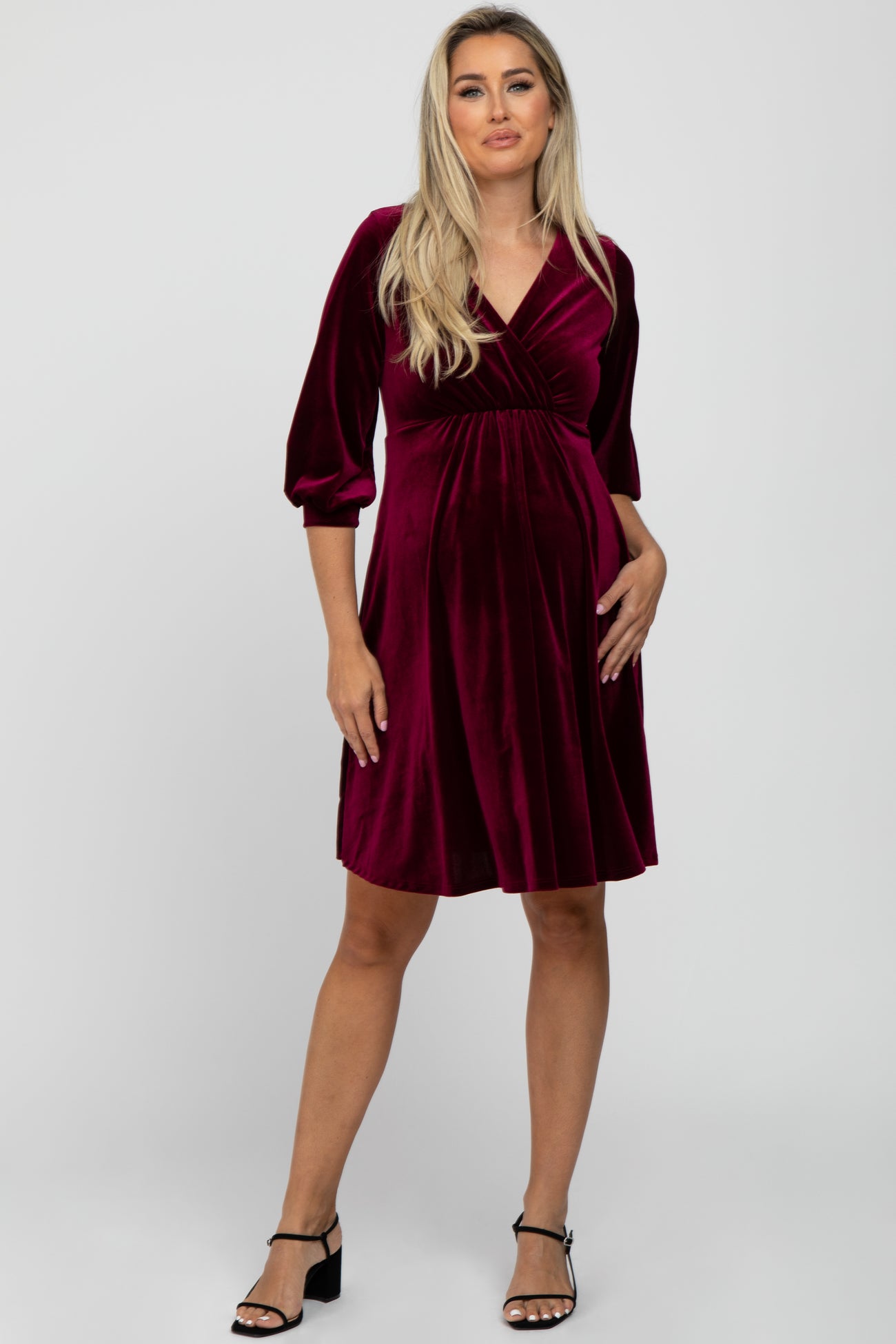woman wearing a burgundy velvet maternity dress with a v-neck and three-quarter sleeves, holding her baby bump