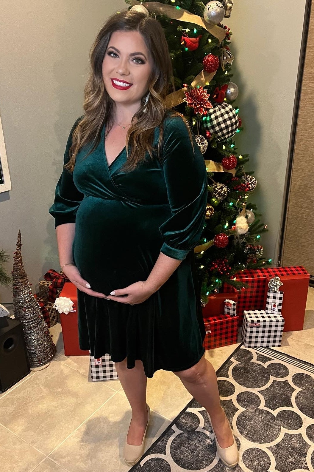 woman wearing a dark green velvet wrap dress, showcasing her baby bump, standing by a decorated Christmas tree