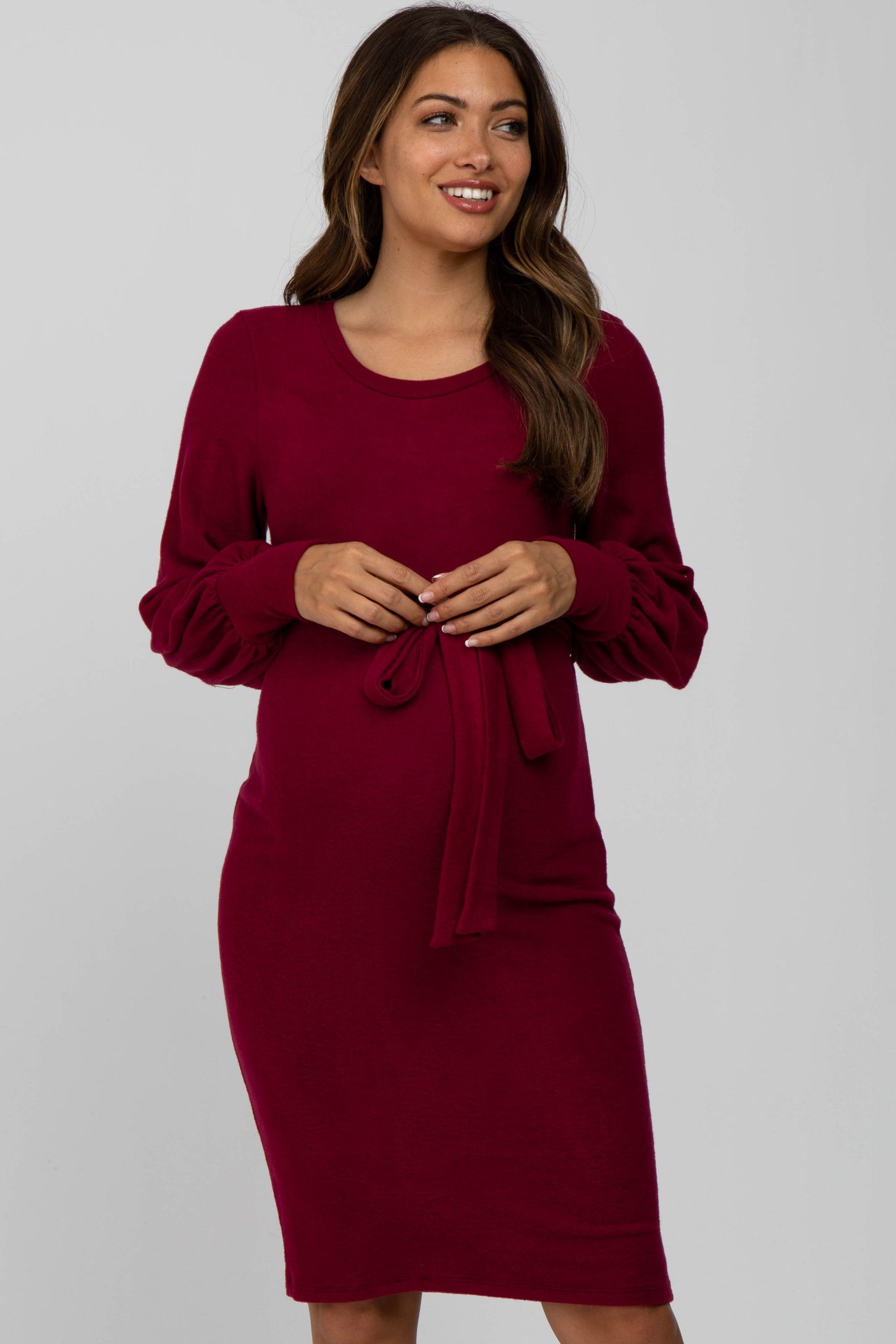 woman wearing a burgundy maternity dress with long sleeves and a tie detail at the waist, showing her baby bump