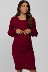 woman wearing a burgundy maternity dress with long sleeves and a tie detail at the waist, showing her baby bump