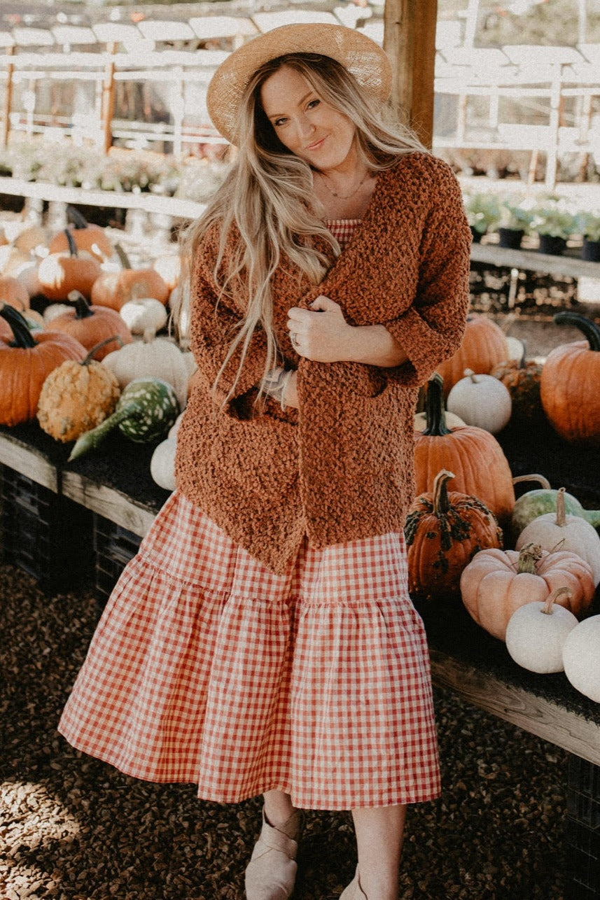 woman wearing a rust-colored oversized cardigan over a red and white gingham tiered dress, standing among pumpkins
