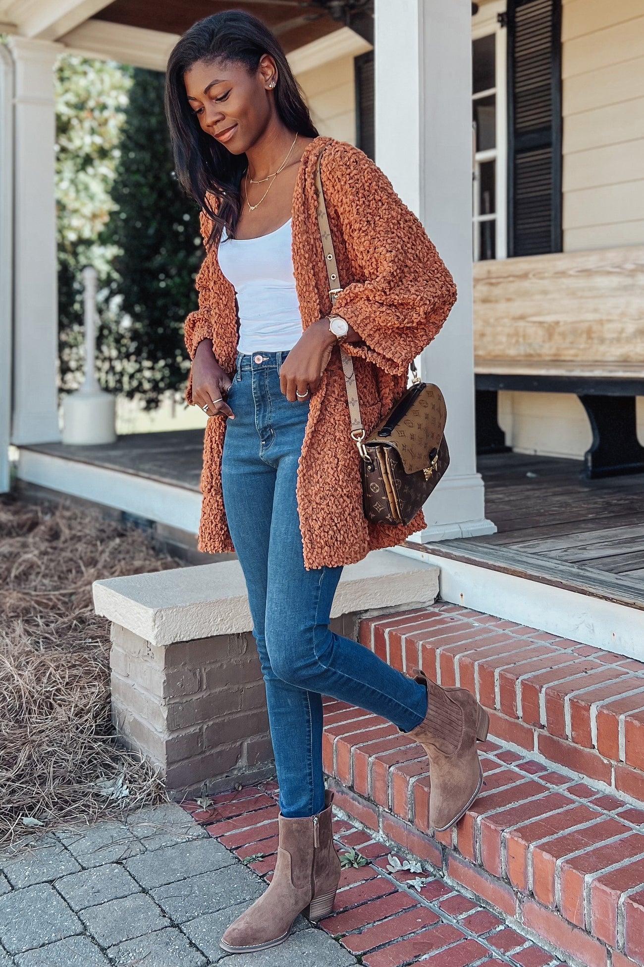 woman wearing a rust-colored fuzzy cardigan, white tank top, and high-waisted skinny jeans with ankle boots and a shoulder bag