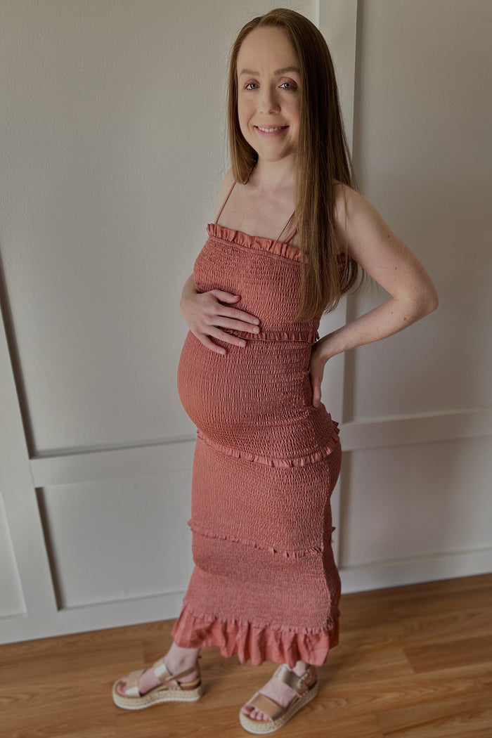 Smiling pregnant woman in a terracotta smocked maternity dress holding her baby bump