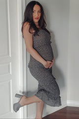 woman wearing a grey floral maternity dress, showing her baby bump while posing by a wall