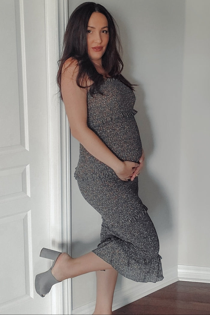 woman wearing a grey floral maternity dress, showing her baby bump while posing by a wall