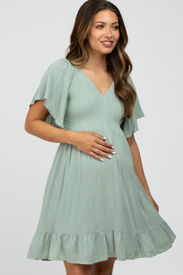 woman wearing a mint green maternity dress with flutter sleeves and a ruffled hem, holding her baby bump