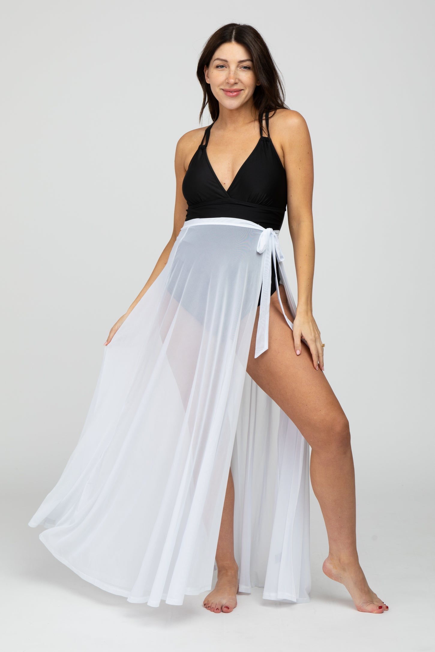 woman wearing a black maternity bikini top and a sheer white maternity cover-up skirt, showcasing a stylish swim look