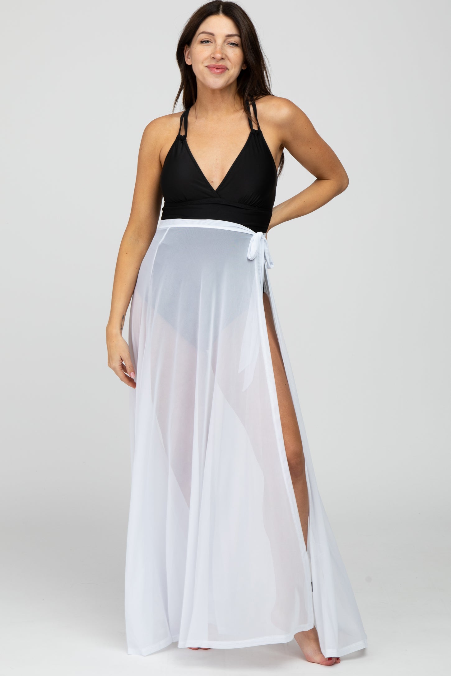 woman wearing a black maternity swimsuit with a white sheer cover-up, smiling and standing with one hand on her hip