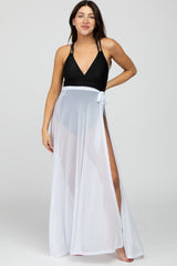 woman wearing a black maternity swimsuit with a white sheer cover-up, smiling and standing with one hand on her hip
