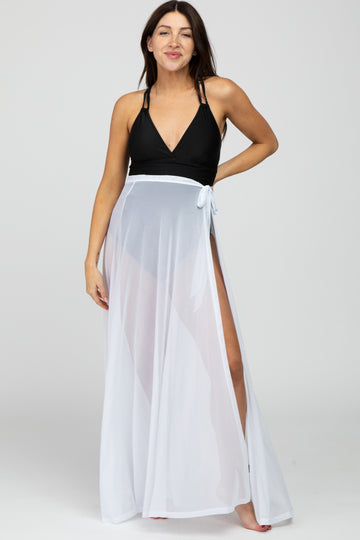 woman wearing a black maternity swimsuit with a white sheer cover-up, smiling and standing with one hand on her hip
