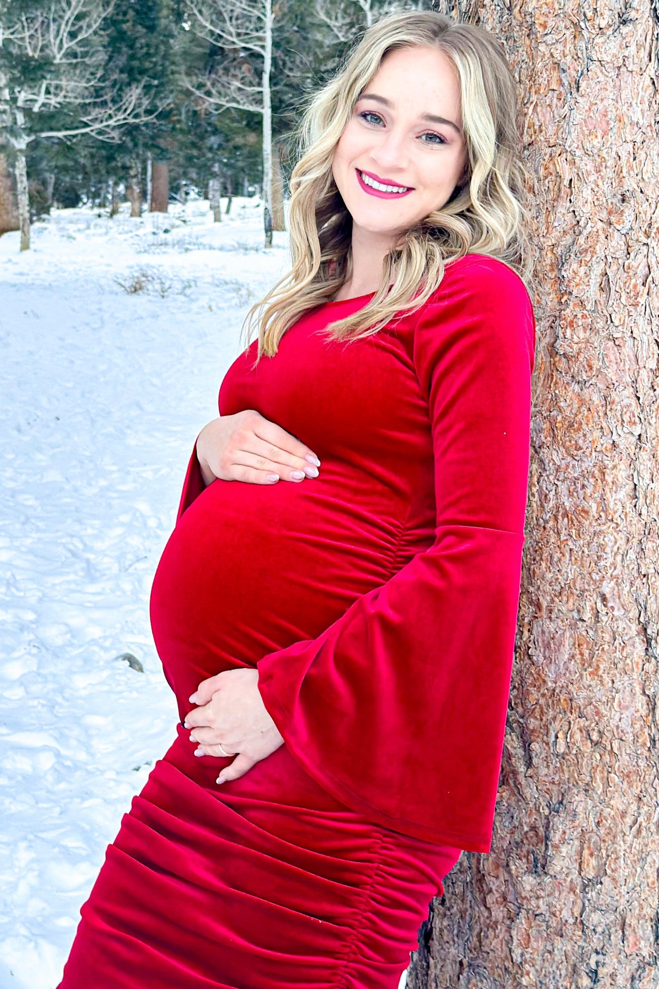 woman wearing a red velvet long sleeve maternity dress, showing her baby bump while leaning against a tree in snow
