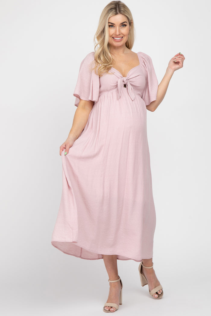 woman wearing a light pink maternity maxi dress with short flutter sleeves and a tie front, showing her baby bump