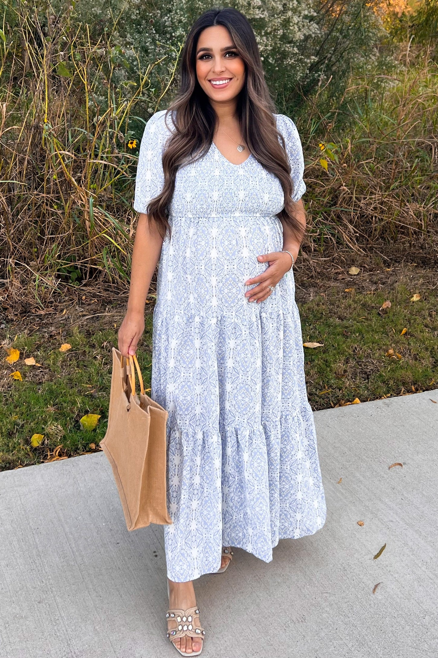 woman wearing a light blue floral maternity maxi dress and holding her baby bump while carrying a tan tote bag