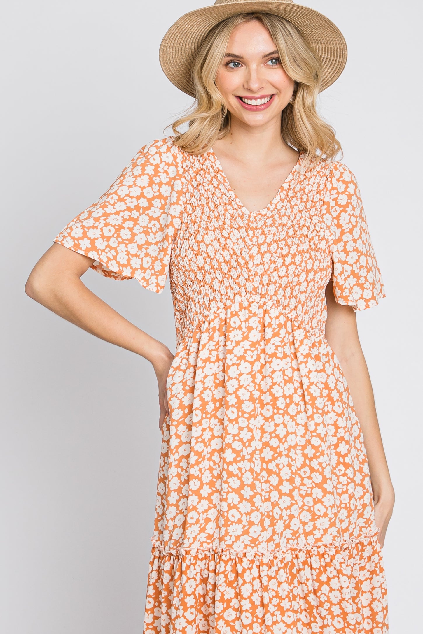 woman wearing an orange floral print short sleeve dress with a fitted bodice and tiered skirt, smiling and wearing a straw hat