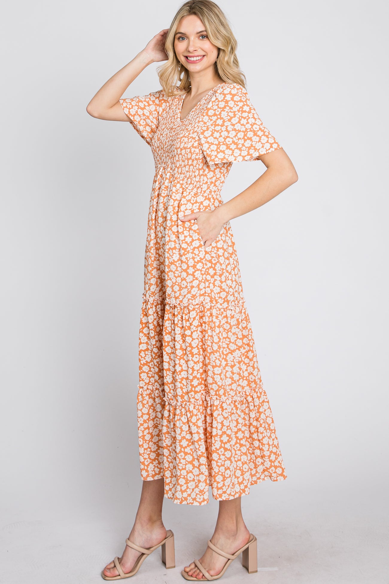 woman wearing an orange floral maxi dress with a v-neckline and short sleeves, standing with one hand in her hair
