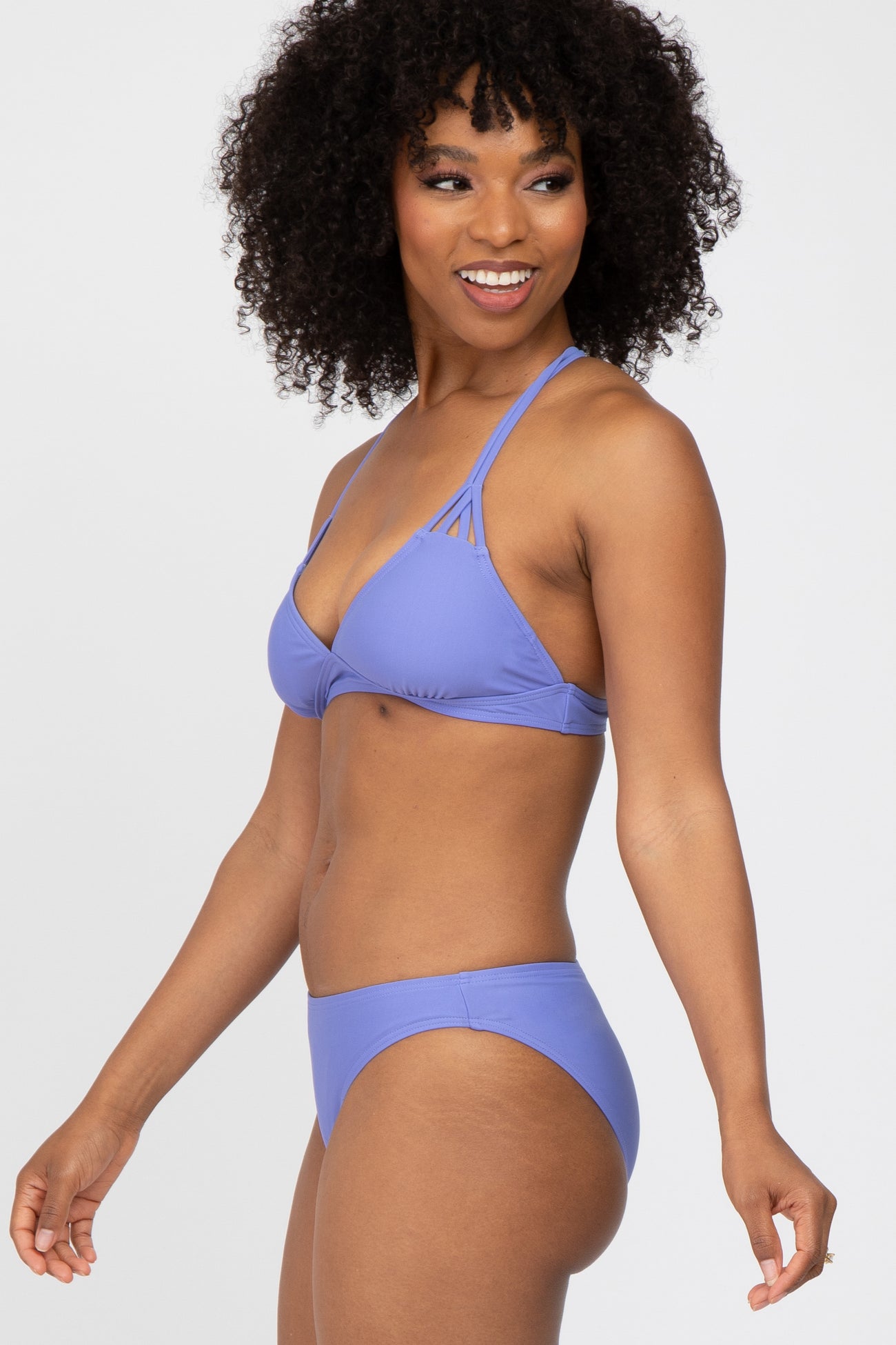 woman wearing a lavender bikini with a triangle top and minimal coverage bottoms, smiling and posing to the side