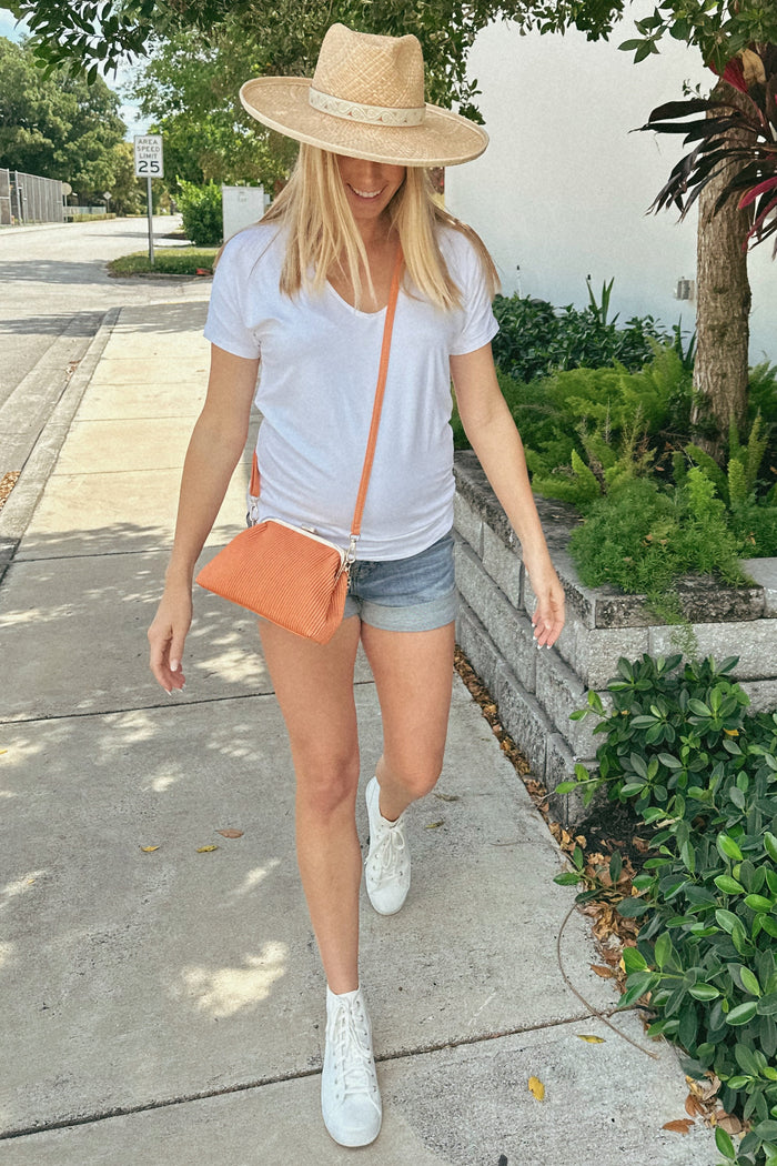 woman wearing a white v-neck maternity top and denim shorts, walking outdoors with a straw hat and orange crossbody bag