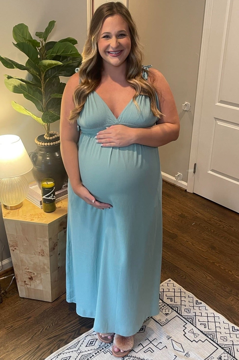 woman wearing a light blue maternity maxi dress, gently holding her baby bump while standing indoors
