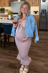woman wearing a pink smocked maternity dress with a bow, blue denim jacket, and holding a drink, showcasing her baby bump