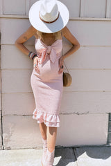 woman wearing a pink maternity dress with a tie front detail and ruffle hem, showing her baby bump and accessorized with a hat.