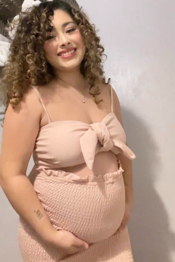 smiling pregnant woman with curly hair wearing a pink two-piece maternity outfit holding her baby bump