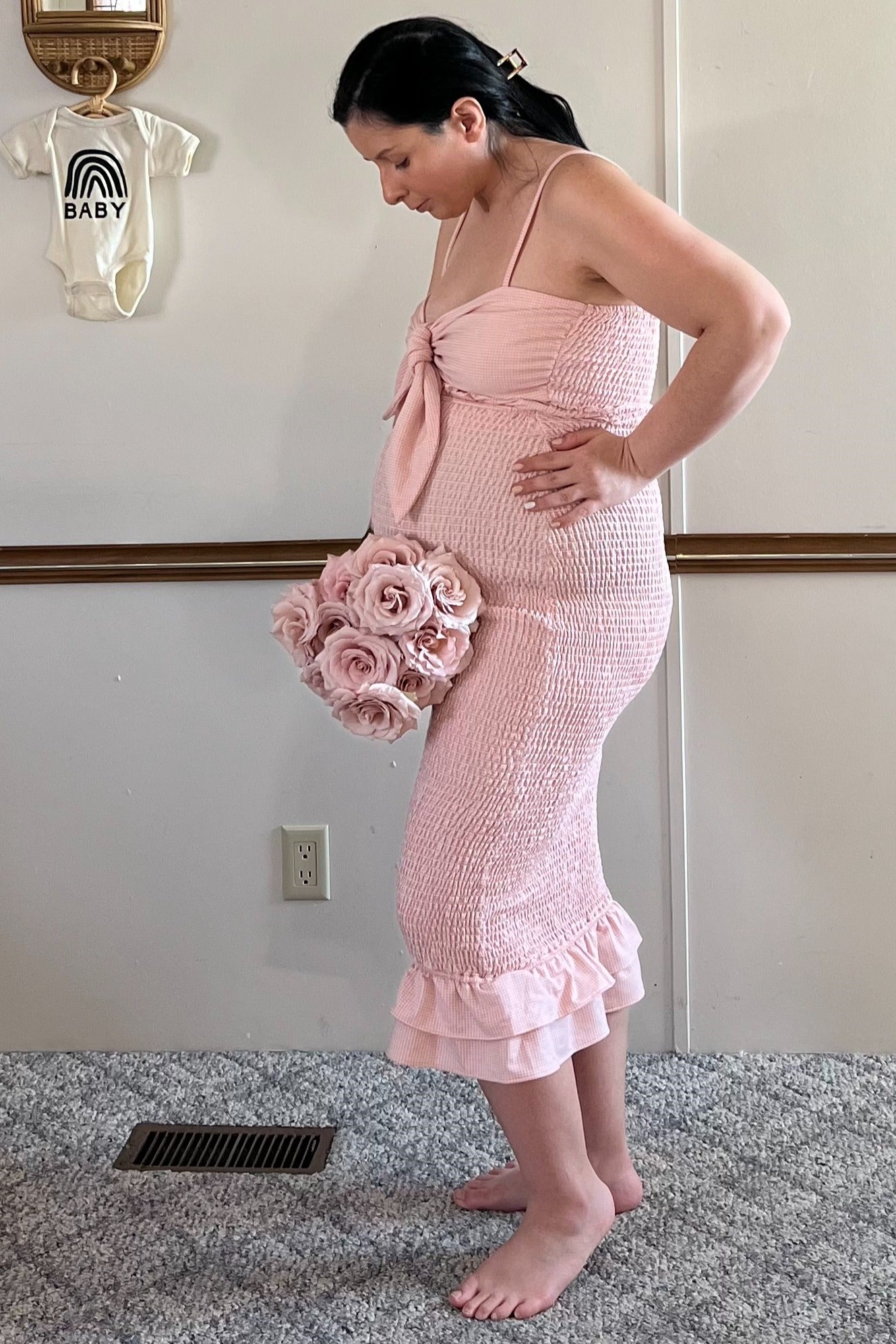 woman wearing a pink smocked maternity dress with a tie front, holding a bouquet of roses and showing her baby bump