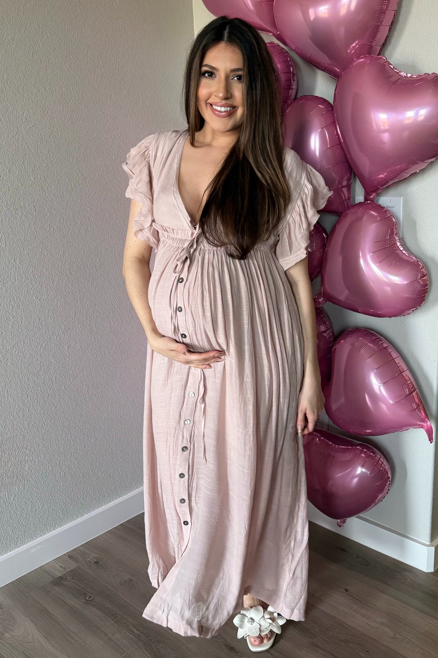 woman wearing a light pink maternity maxi dress with button details and holding her baby bump, with heart balloons in the background