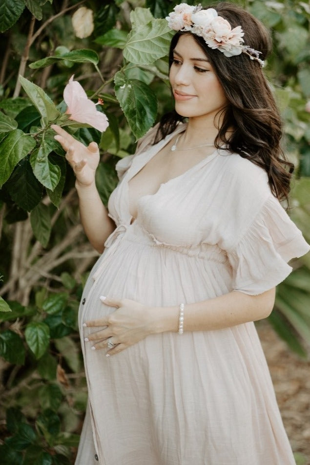 woman wearing a soft pink maternity dress with flutter sleeves and a baby bump, reaching out to a butterfly in a garden