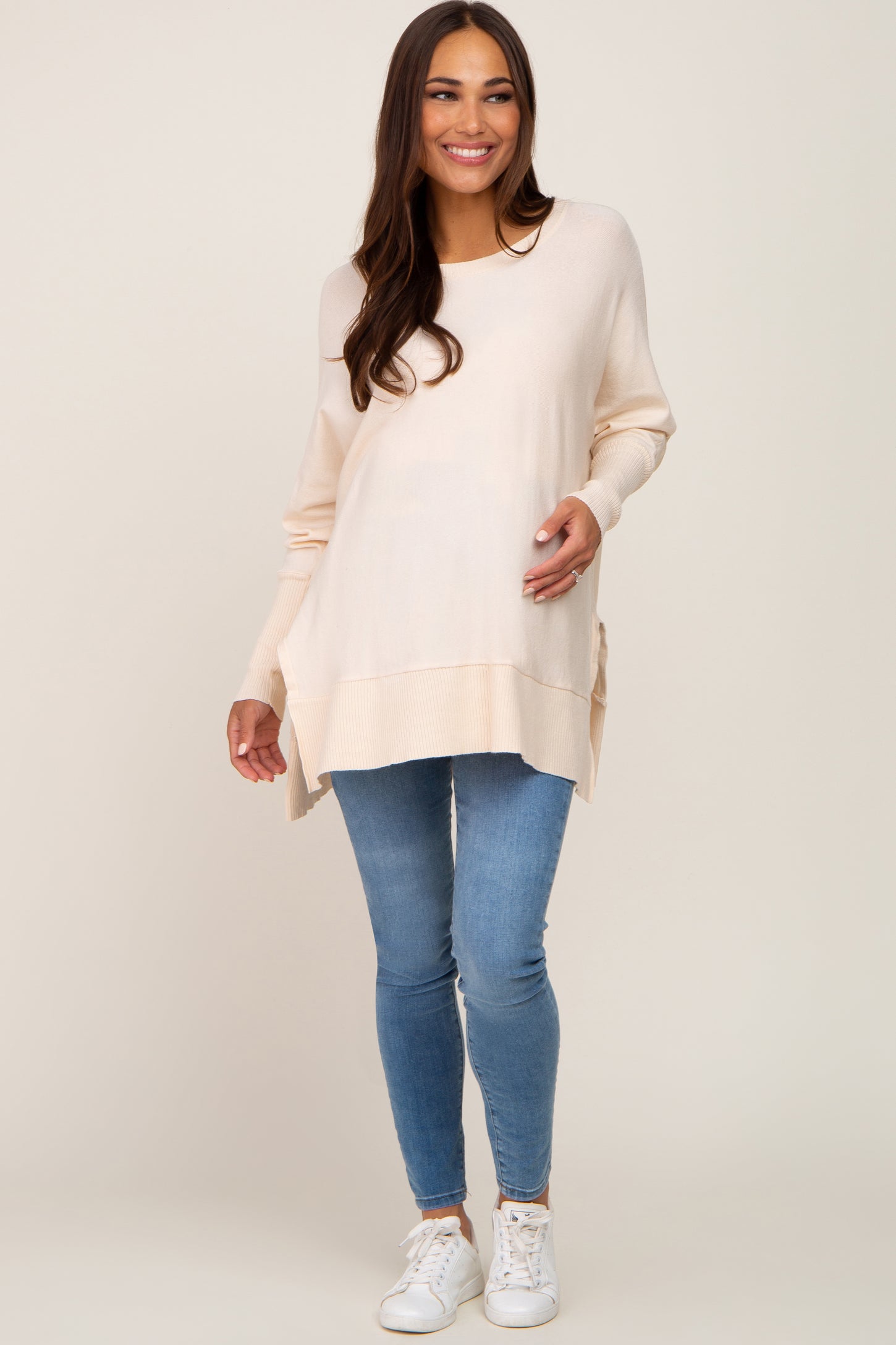 woman wearing a light beige maternity sweater with side slits and skinny jeans, holding her baby bump