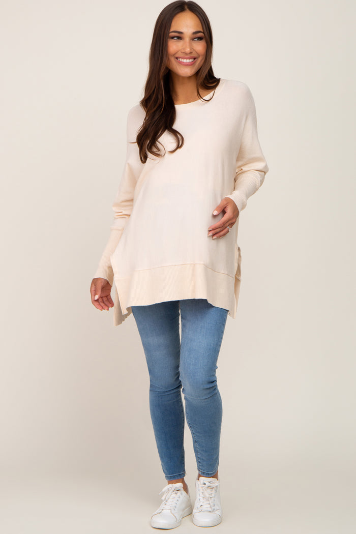 woman wearing a light beige maternity sweater with side slits and skinny jeans, holding her baby bump