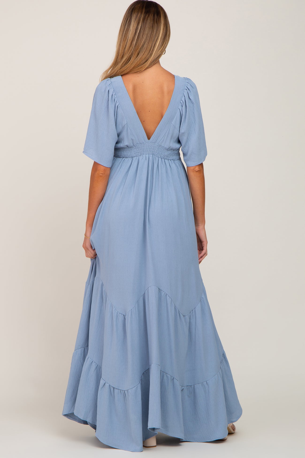 woman wearing a light blue maternity maxi dress with a smocked waist and tiered ruffles, showing a deep V-back