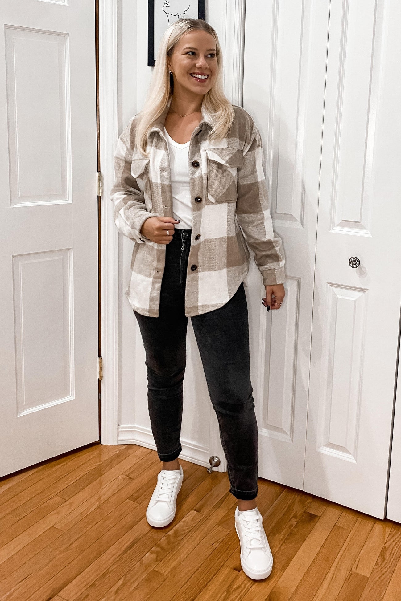 woman wearing a tan and white plaid shacket with black jeans and white sneakers standing in front of a white door