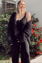 smiling woman wearing a black jumpsuit and chunky knit cardigan standing in front of red flowers