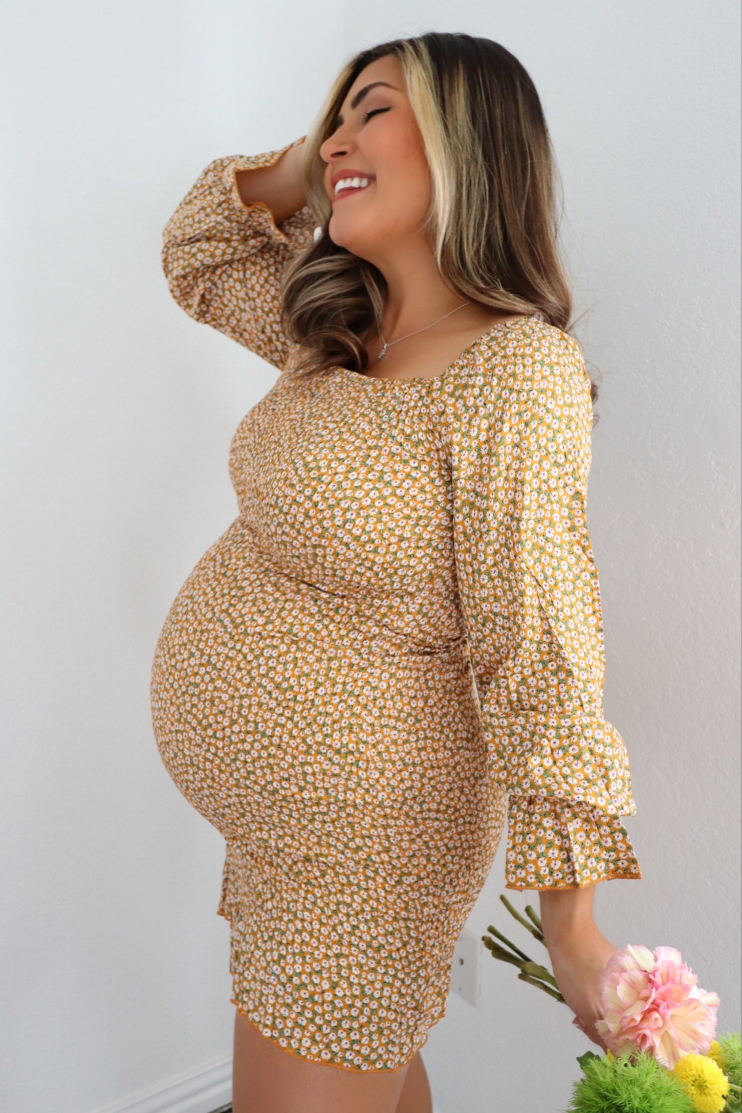 woman wearing a mustard yellow floral maternity dress and holding flowers, with her baby bump visible