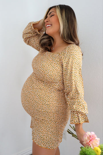 woman wearing a mustard yellow floral maternity dress and holding flowers, with her baby bump visible