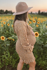 woman wearing a brown floral maternity dress holding her baby bump in a sunflower field at sunset