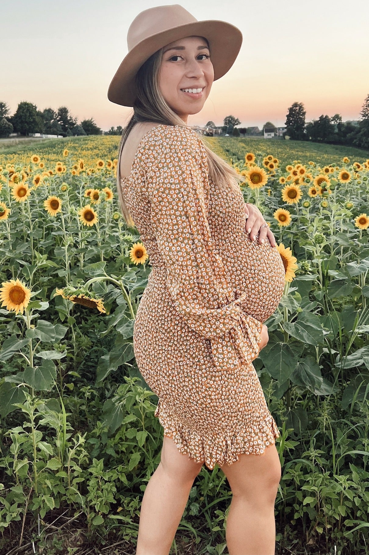 woman wearing a floral print maternity dress with ruffle details and a baby bump, standing in a sunflower field