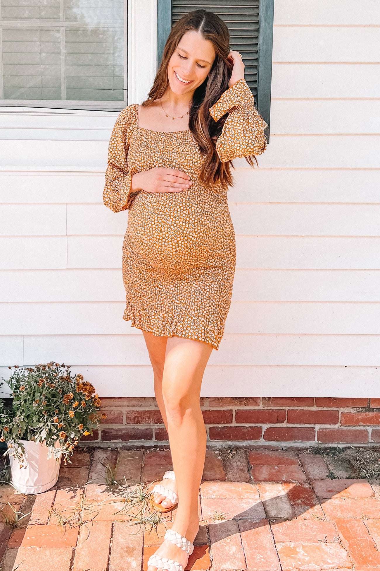 woman wearing a mustard yellow floral maternity dress and holding her baby bump outside a house