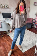 woman wearing a taupe striped button-up shirt and blue jeans, holding a pink phone, standing in a home office setting