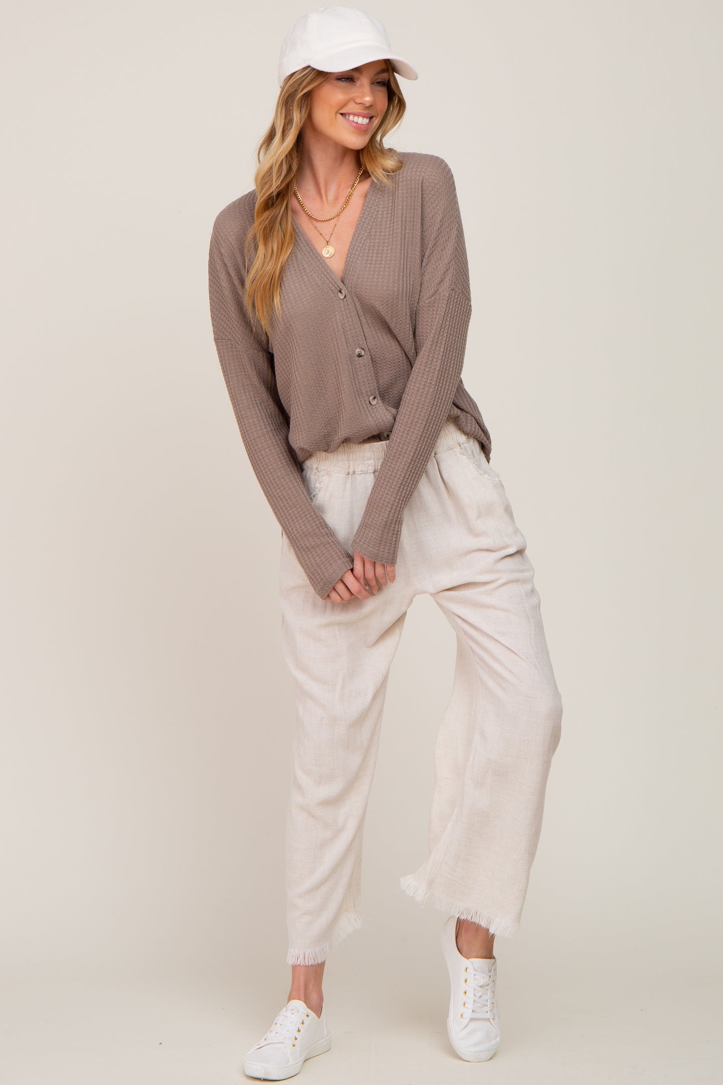 woman wearing a taupe button-up ribbed cardigan, cream pants, and a white cap, styled casually with white sneakers