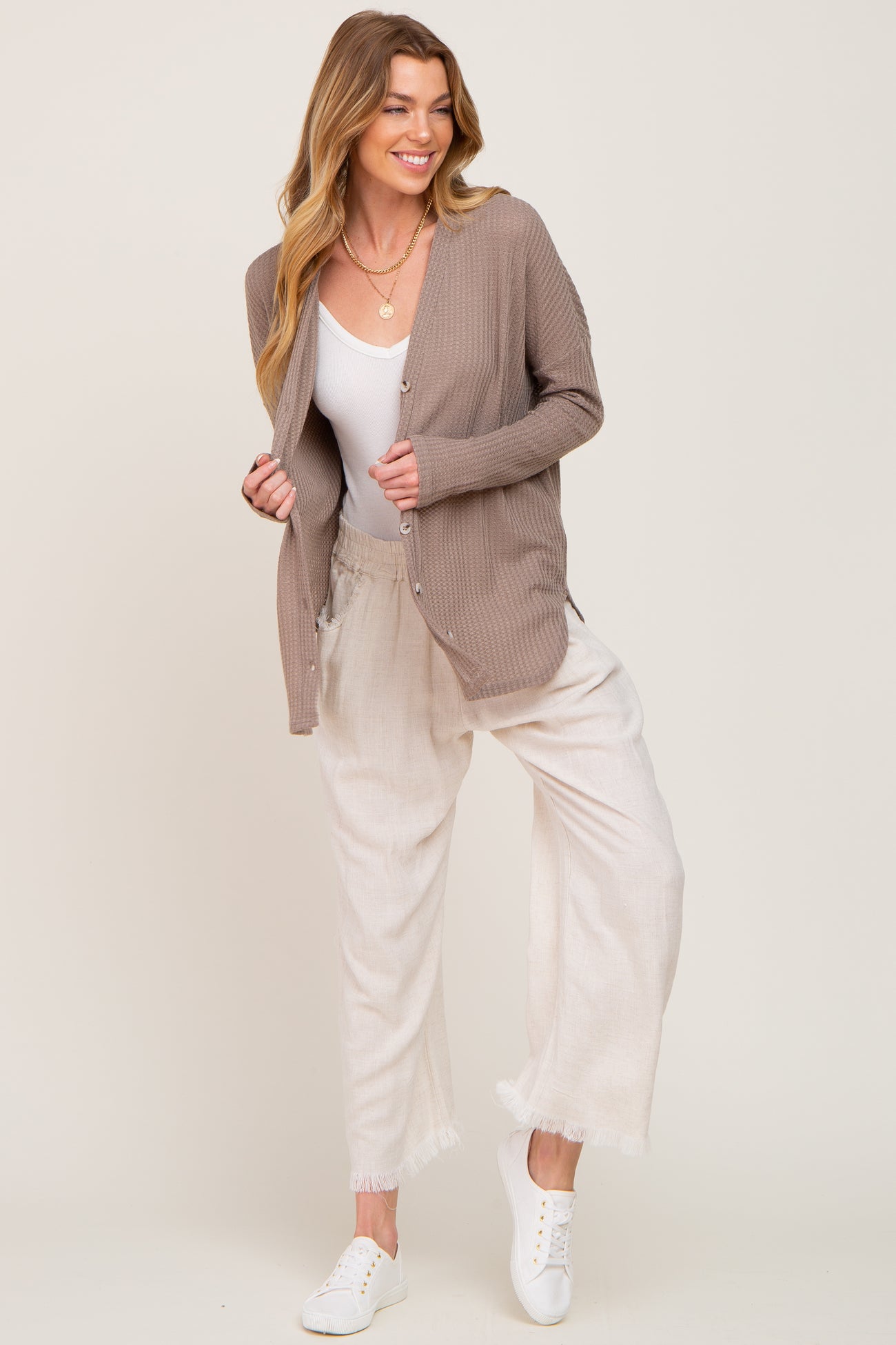 woman wearing a beige ribbed cardigan over a white tank top and light-colored loose-fitting pants with frayed hems