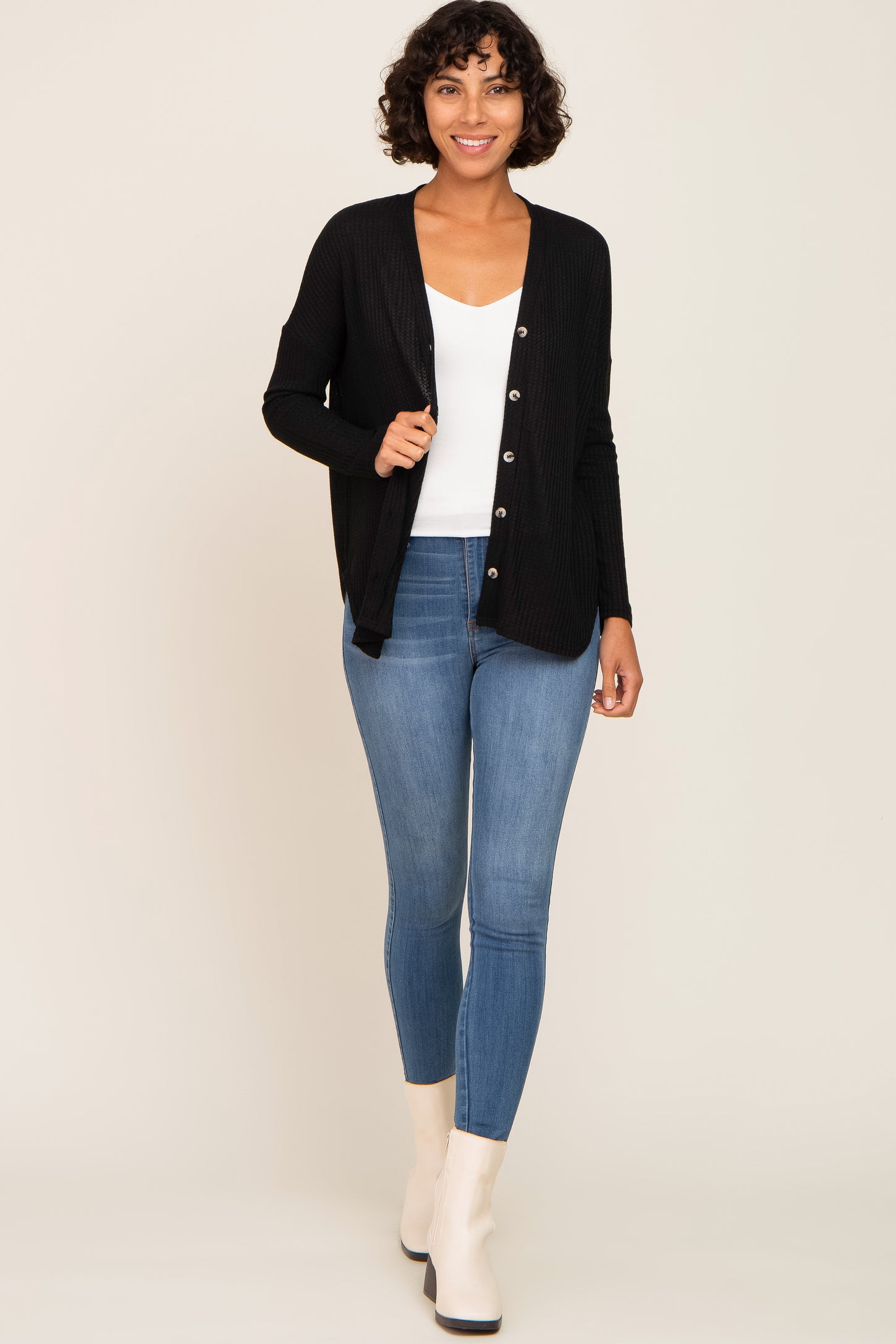 woman wearing a black ribbed cardigan over a white top and blue skinny jeans, paired with white ankle boots