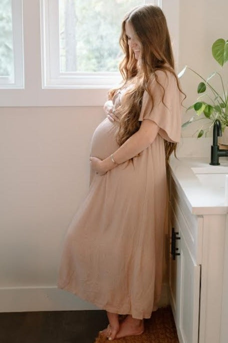 woman wearing a beige maternity maxi dress and showing her baby bump while standing near a window