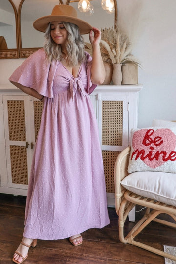 woman wearing a light pink polka dot maternity maxi dress with tie front and flutter sleeves, accessorized with a wide-brim hat