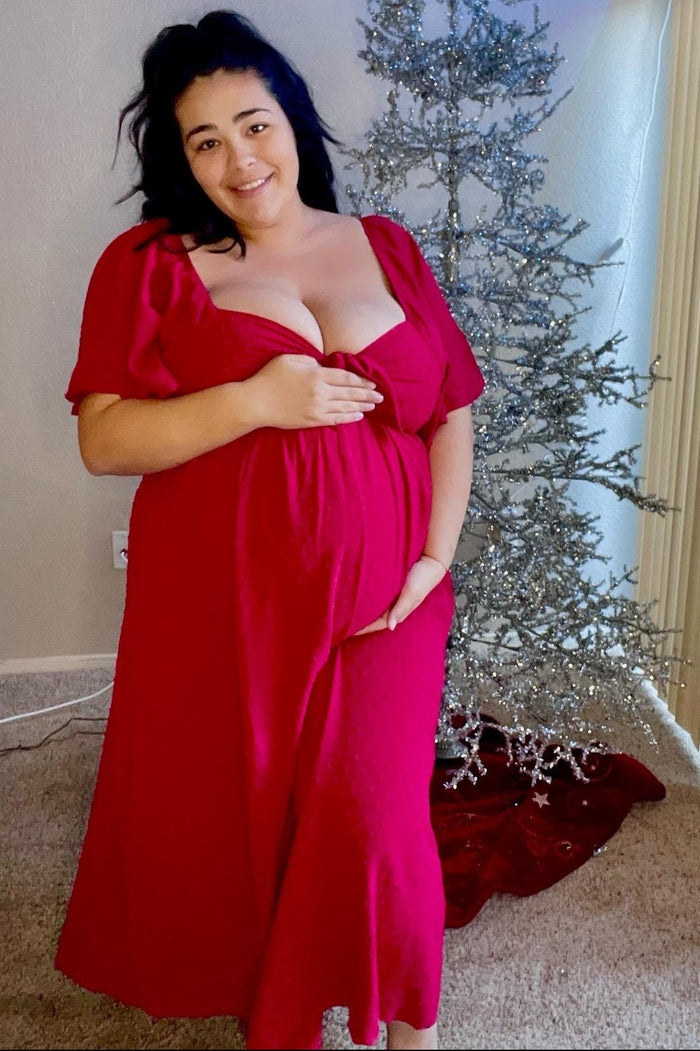 woman wearing a bright red maternity dress with short sleeves and a baby bump, standing next to a beautifully decorated Christmas tree