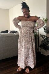 woman wearing a floral off-the-shoulder maxi dress with long sleeves and a flowy skirt, posing with a smile indoors