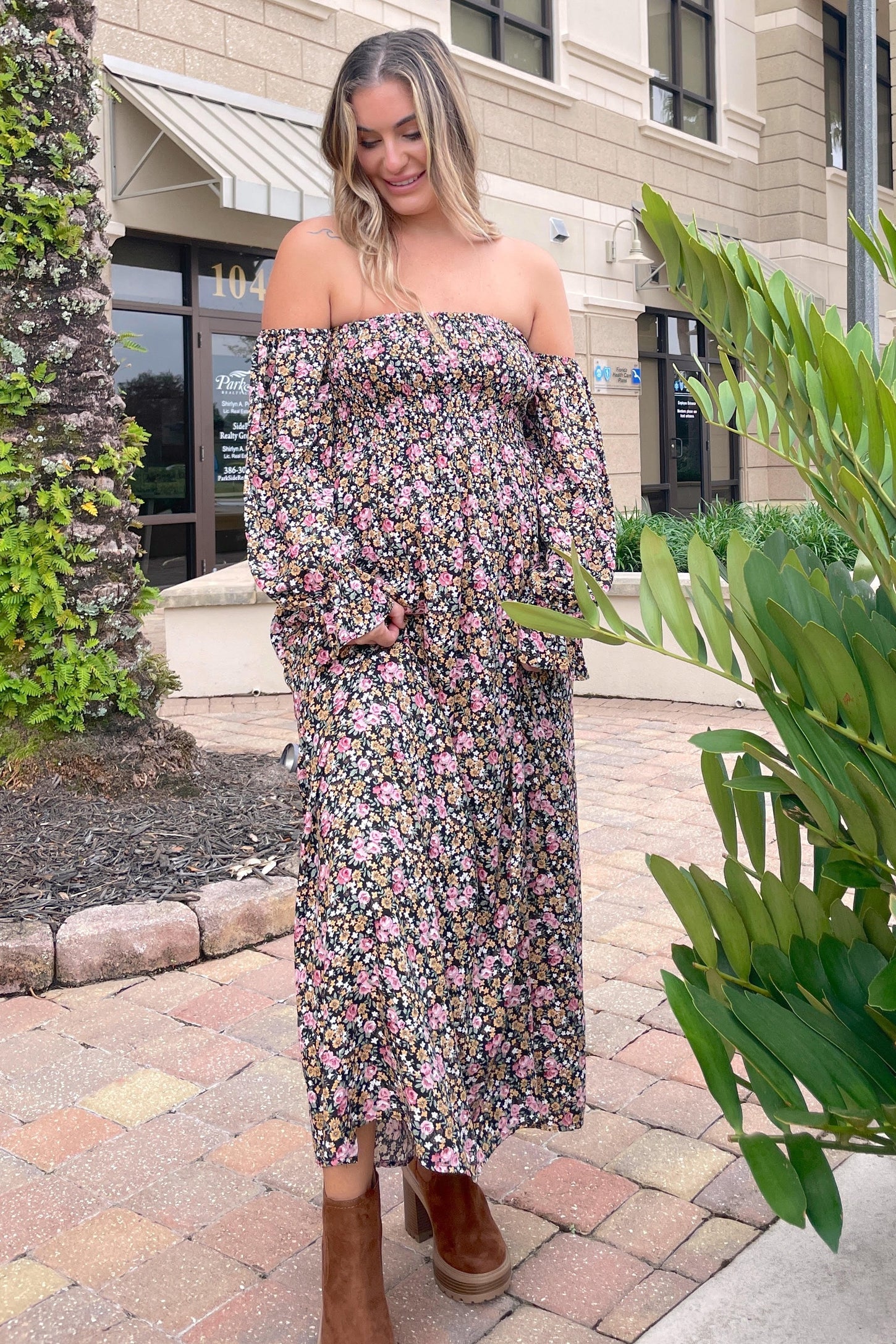 woman wearing a floral off-the-shoulder maternity maxi dress with a baby bump, standing outside with greenery around
