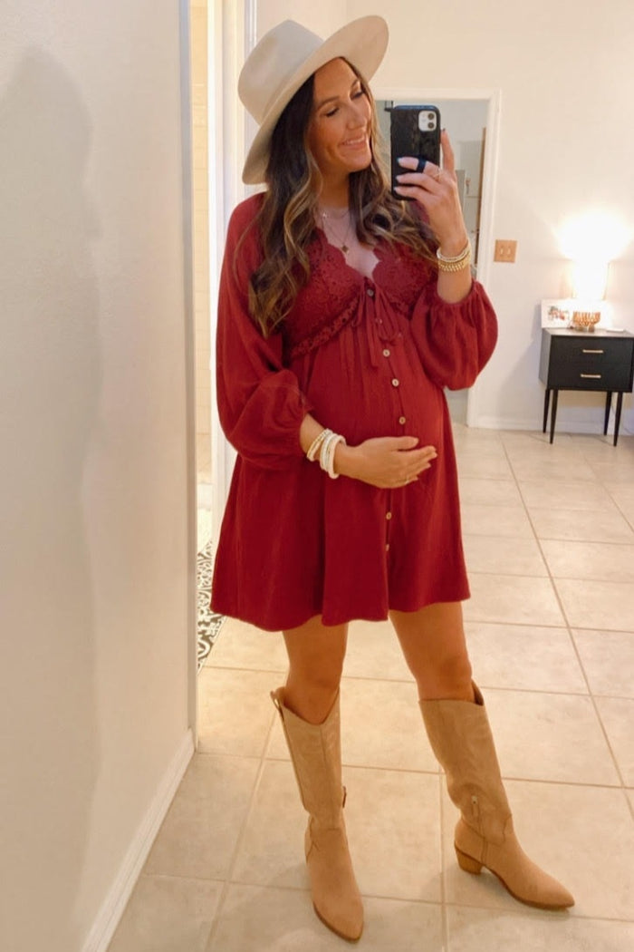 woman wearing a burgundy maternity mini dress with buttons and bell sleeves, holding her baby bump, paired with tan knee-high boots and a wide-brimmed hat
