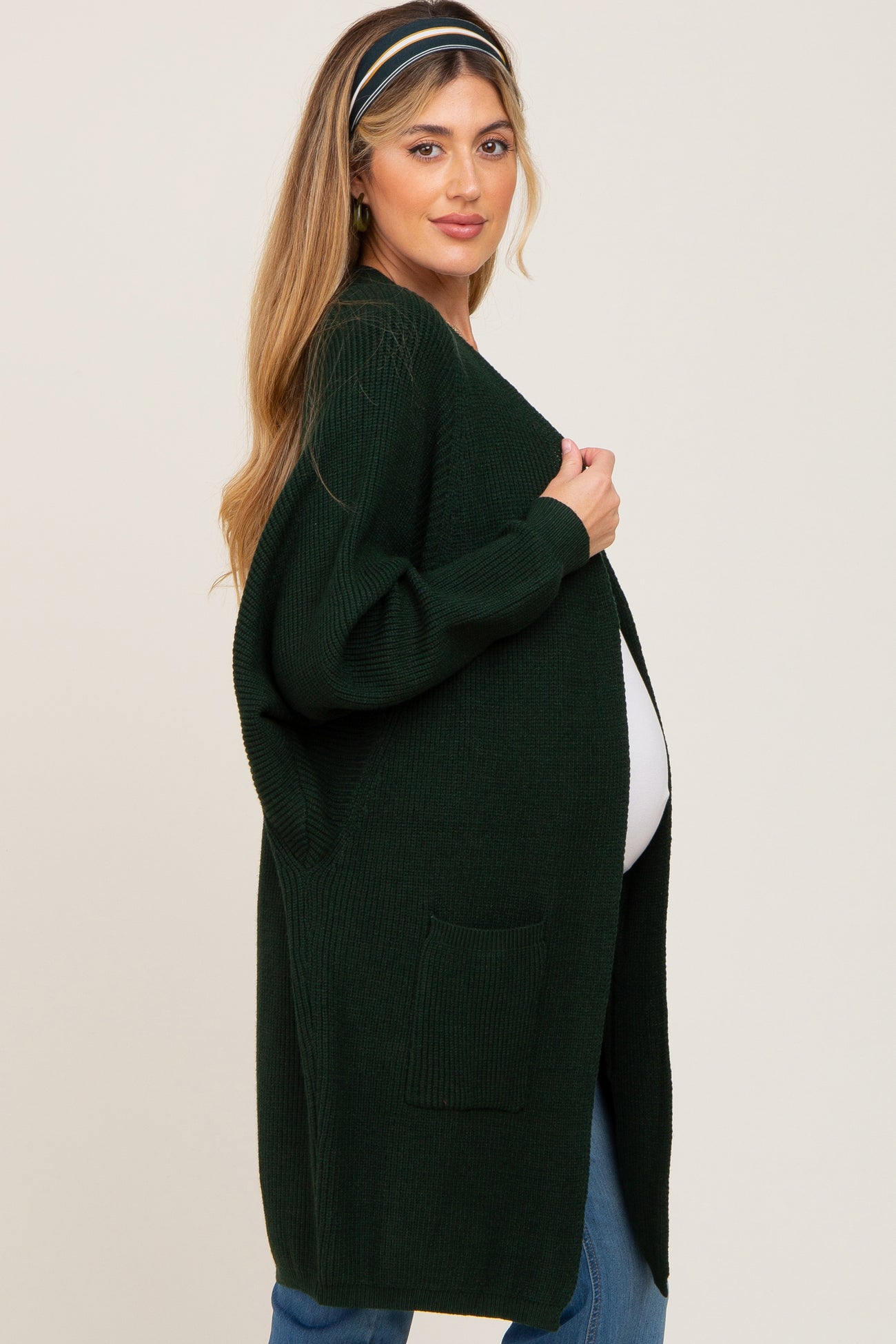 woman wearing a dark green maternity cardigan, standing sideways to show off her baby bump and holding the cardigan open
